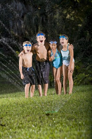 Four happy kids standing arm in arm shouting and laughing, soaked by lawn sprinklerの写真素材