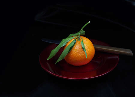 Mandarin type small orange on red plate, with leaves. Whole, unpeeled. With knife, dark background.の写真素材