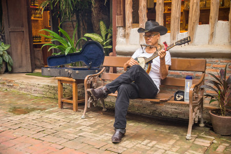 RATCHABURI, THAILAND - JUNE 10, 2017: Senior man sitting on the bench in fron of his shop playing...