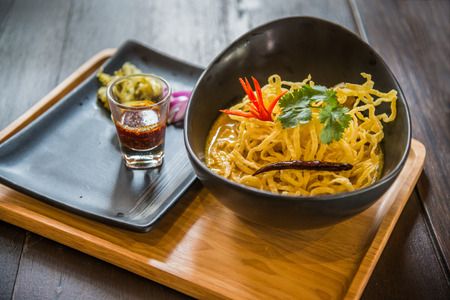 Thai northern traditional style noodle in curry with red onion, pickle and a glass of oil chili on wooden plate on tableの写真素材