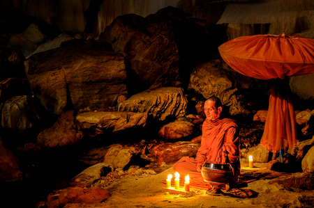 Kanchanaburi, Thailand - July 15, 2011: Buddhist monk sitting with alms bowl, lighten candle and long-handles umbrella to do individual meditation in the cave in national park in Kanchanaburi, Thailandのeditorial素材