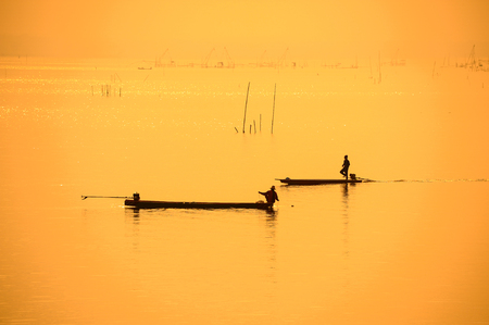 Fishermen on boat in silhouette using fishing net to fish in rural swamp during sunsetの写真素材