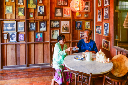 Krabi, Thailand - May 3, 2015: Senior man and senior woman drinking tea together on white marble table in traditional wooden house where the wall decorated with people's pictures in Krabi, Thailandのeditorial素材