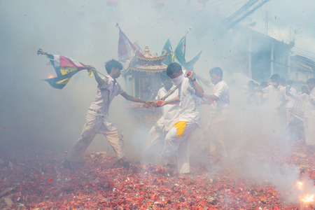 Phang Nga, Thailand - October 14, 2018: Group of men in white dress holding palanquin with Chinese god statue inside  marching on street while people fire firecracker to worship the god in vegetarian festival parade in Phang Nga, Thailandのeditorial素材