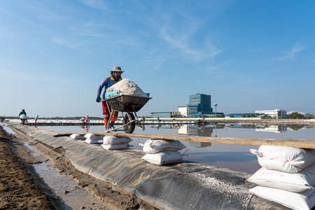 Samutsongkram, Thailand - March 10, 2019: Salt farmer is puching wheelbarrow full of salt on salt farm in Samutsongkram, Thailandのeditorial素材