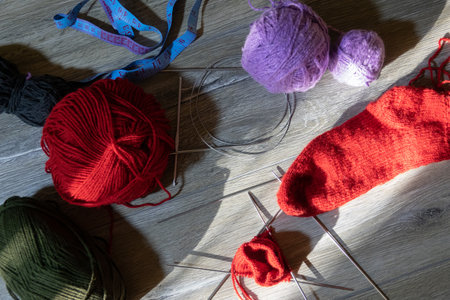 Sock in the process of knitting with needles and yarn of red color on a gray wooden table. Clews threads of red, lilac, dark green, and black color on a gray wooden table. View from aboveの写真素材