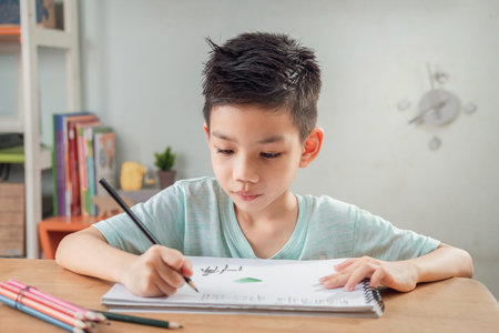 Portrait of little Asian Boy Drawing with a Pencil while Sitting Behind School Desk Home.の写真素材
