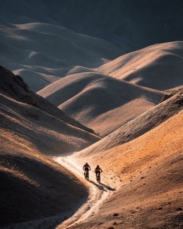 A Two cyclists ride through winding trail in sunlit desert landscape, surrounded by rolling hills and natural tonesの素材