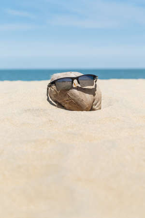 Sunglasses over coconut fruit on sandy beach under hot shiny sun light looked like a happy (tourist) man lying on the beach during holiday summer vacation on wonderful trip under bright blue sky.の写真素材