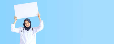 Young Arab Muslim doctor women smile showing white card on wide blue background concept for Islam people working in medical hospital health care, Nurse wear hijab in pandemic stay home panoramic text.の写真素材