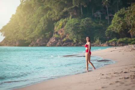 Young beauty asian woman in dress happy travel smile walk on beach sea near blue water wave for trip vacation. Summer day tan skin girl happy in red bikini smiling at sunbath on koh chang, Thailand.の写真素材