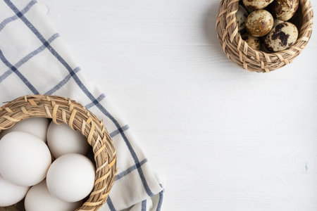 White chicken, quail eggs in baskets and a towel on a white wooden background with space for text....