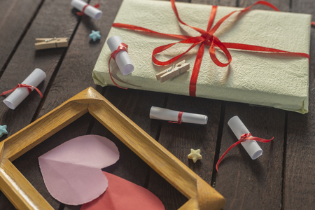 Flat lay romantic background. Top view to giftbox, rolled wish papers, and wooden frame with paper hearts on dark wood board.の写真素材