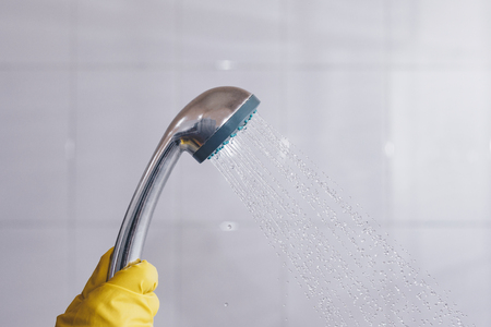 A person in rubber gloves holding showerhead with flowing water. Women in yellow protective gloves holding the head of the shower with running water in the bathroomの写真素材