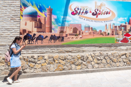 BUKHARA, UZBEKISTAN - MAY 25, 2018: Silk and Spices Festival 2018. School girl with mobile phone walking against the banner of the festival in Bukhara, Uzbekistanのeditorial素材