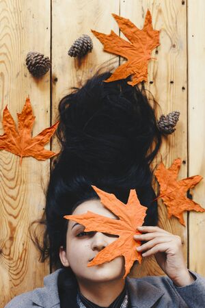 Autumn woman. Fall season concept. A portrait of lying on wooden floor woman. Young, beautiful and happy female holding a big yellow autumn leaf. Pretty girl hiding her face with fall leafの写真素材