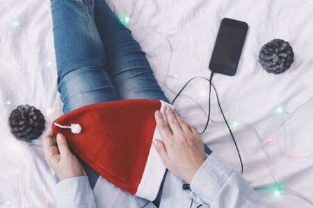 Female listening to music in bed. Pine tree cones, Santa Claus cap and smartphone with earphones near the legs of female. Christmas holidays. X-mas morning in a cosy bedroom.の写真素材
