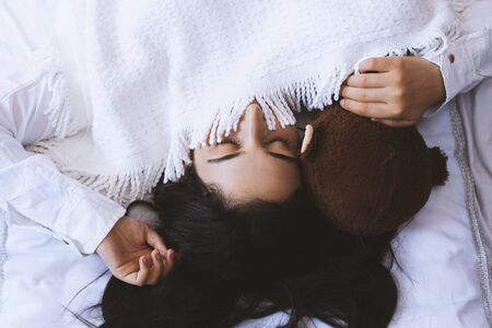 Young woman sleeping with toy bear. Cute black haired woman resting in bed with pleasure. Lonely woman in bed with bear toy. Happy life of single lady. Pretty girl enjoying long sleep on her weekendの写真素材