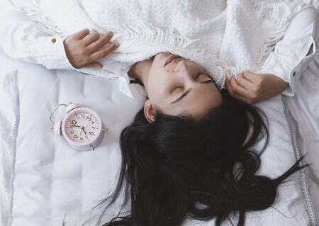 Beautiful black-haired woman sleeping in cozy bed. The alarm clock set at 7 a.m. morning. Female is going to wake up at 7 o'clock early morning. Top view to cute and sweet female.の写真素材