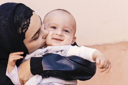 Young Muslim woman in hijab holding her 1-year-old baby boy, hugging and kissing. Happy Muslim mother in abaya clothing kiss and play with her little child. Cute family, middle-eastern mother and sonの写真素材