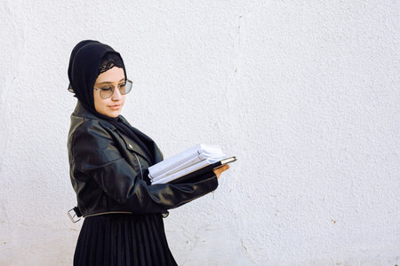 Young middle-eastern student girl holding in hands stack of notepads and her handbag. Modern Muslim...