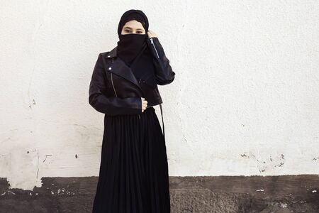 Beautiful Iranian girl in black dress and leather jacket. Attractive Muslim woman in hijab covering her face with a black scarf. Middle-eastern female in traditional Islamic clothingの写真素材
