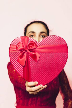 Portrait of lovely woman covering face with giftbox. Valentines day gifts. Beautiful middle-eastern girl receives heart-shaped present box. Love, anniversary, birthday celebration. Feel happy and shyの写真素材