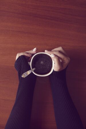 Woman in coffee shop. An unrecognizable woman drinking coffee at cozy home environment. Female hands holding cup of coffee on wooden table backgroundの写真素材