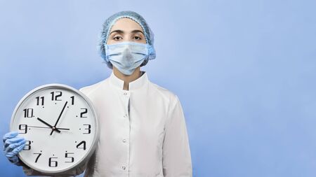 Female doctor in medical protective mask and gloves holding a big clock.の写真素材