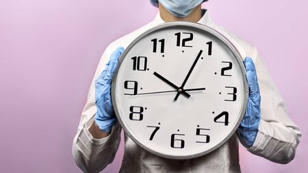 Female doctor in medical protective mask and gloves holding a big clock.の写真素材