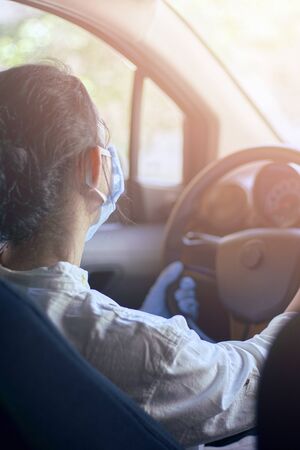 A young woman in a surgical face mask and gloves driving the car during coronavirus pandemic. A female driver in protective gear, preventing infection of covid-19 virusの写真素材