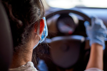 A young woman in a surgical face mask and gloves driving the car during coronavirus pandemic. A...