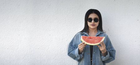 Portrait of a beautiful young lady eating watermelon at outdoors. A little girl in trendy denim and sunglasses holding in hands big slice of sweet fresh juicy watermelon. Face expressionの写真素材