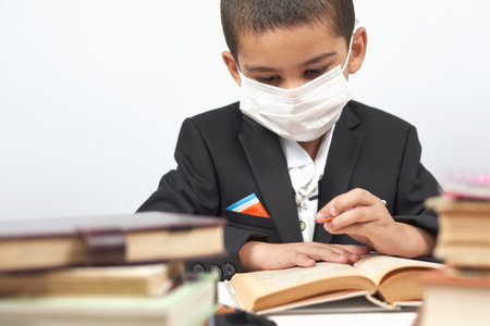 Young schoolboy in face mask doing homework between pile of books. Little kid in black school wear...