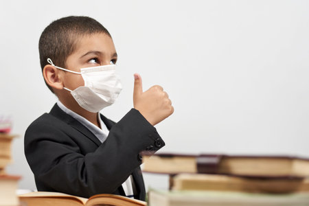 Happy schoolboy in face mask. Mixed-race kid shows thumbs-up gesture between pile of books. Back to...