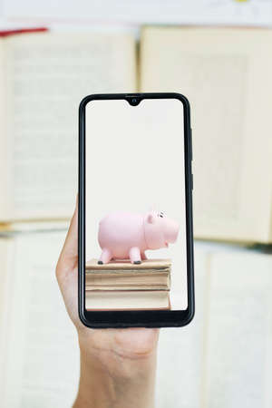 Cost of online education. Person hold smartphone with image of piggy bank and books. Copy spaceの写真素材