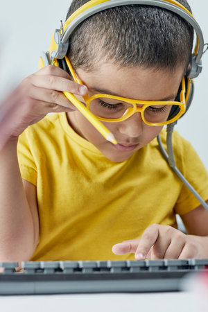 An adorable kid in a yellow t-shirt with a headphone listening online course. Boy typing on pc...