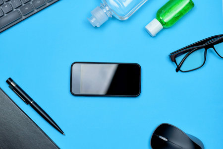 A mobile phone, bottles of hand sanitizers, eyeglasses, pc mouse, keyboard and notepad on blue...