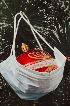 Celebrating winter and Christmas holidays in the new Covid-19 reality. Christmas toy hanging on the Xmas tree with a medical face mask. Merry healthy Christmasの写真素材