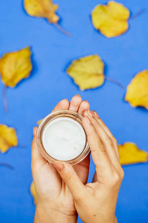 Autumn cream over fall leaves. Cropped shot of female hands holding a facial moisturizing cream over bright colorful autumn background with orange tree leavesの写真素材