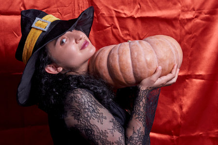 Halloween lady wearing witch cup and holding a big Halloween pumpkin. A woman in witch hat and Halloween pumpkin over orange background. Portrait of young female in traditional black witch capの写真素材