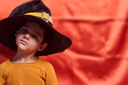 A young kid in orange sweater wearing witch hat. Portrait of 8 years old boy with a big black halloween capの写真素材