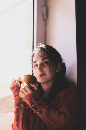 Cold autumn days - a young multi-racial female drinks coffee in a cozy windowsill. Middle-eastern or...