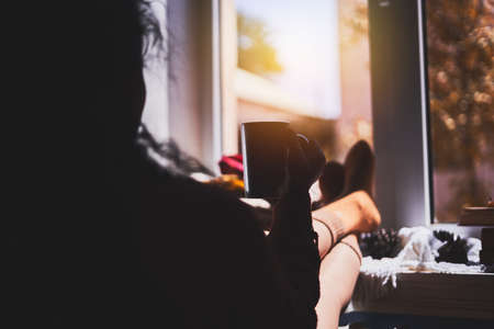 A cozy autumnal day. A woman hanging her legs on the window sill. Cold autumn sunny day. Drink coffee near the windowの写真素材