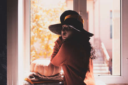 Halloween lady wearing witch cup. A woman in witch hat and Halloween pumpkin near the windows. Portrait of young female in orange sweater and traditional black witch capの写真素材