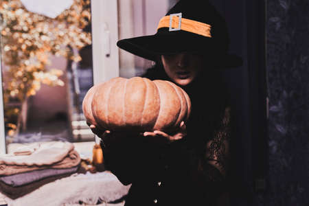 Halloween lady wearing witch cup. A woman in witch hat and Halloween pumpkin near the windows. Portrait of young female in orange sweater and traditional black witch capの写真素材