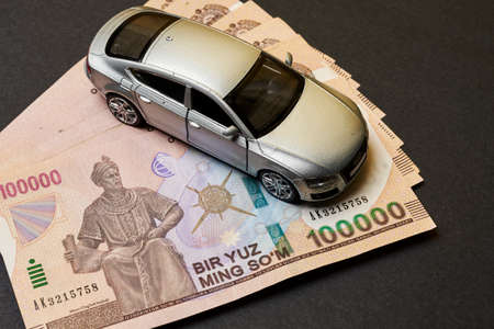 Uzbek sums and car toy. Concept of auto vehicle insuarance in Uzbekistan. Pile of 100000 sums and modern automobile toy on black background. Auto tax, auto insurance, automobile tax conceptsの写真素材