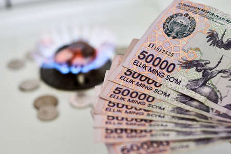 Burning gas stove burner and Uzbek money - sum banknotes. Pay for natural gas, bill, tariff. Growing tariffs for gas. Pile of coins and burning fire gas stove hob and moneyの写真素材