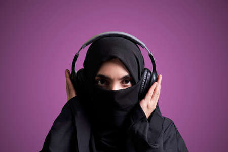 Muslim woman in hijab listening to music with wireless headset. Arabic girl in traditional nikab clothing listening favorite music with wireless headphonesの写真素材