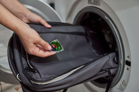 Washing and cleaning backpack in washing machine. Loading dirty school backpack into washing machine. Clean backpack. Washing school bag. Person places backpack in wash machineの写真素材